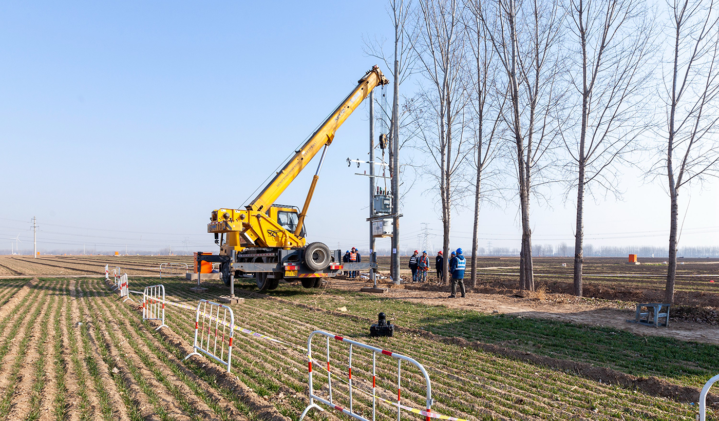 施工现场，工作人员分工协作，依次进行挖坑、立杆、安装金具横担绝缘子及附件、跌落式熔断器、避雷器、变压器及配电箱等系列作业。图3为，2026年1月8日上午，在河南省安阳市滑县半坡店镇杏头村的农田里，杏头5号机井通电配套工程施工现场，吊车正在吊装变压器。（王亚方 摄）